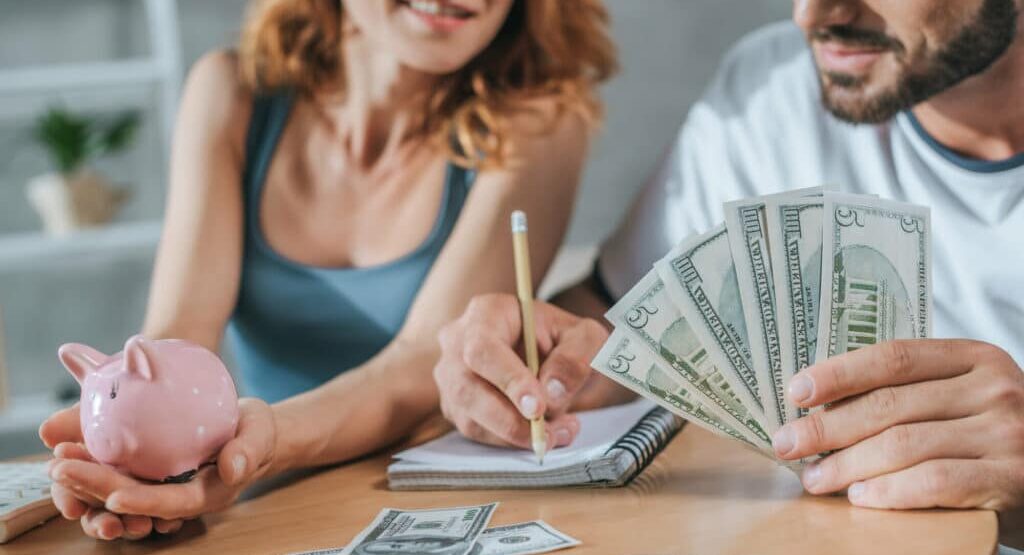 man and women sitting at table holding money and piggy bank