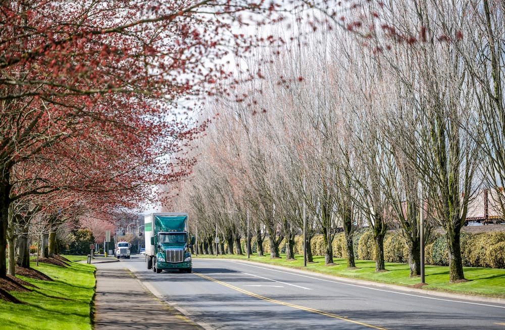 Freight Shipping in the Spring teal semi truck driving down road with budding trees on either side