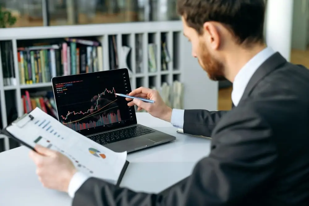Dynamic Pricing businessman looking at graph on laptop with a report in one hand and a pen pointing at laptop screen in the other hand