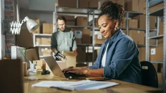 woman typing on computer at desk with man packaging box in back preview