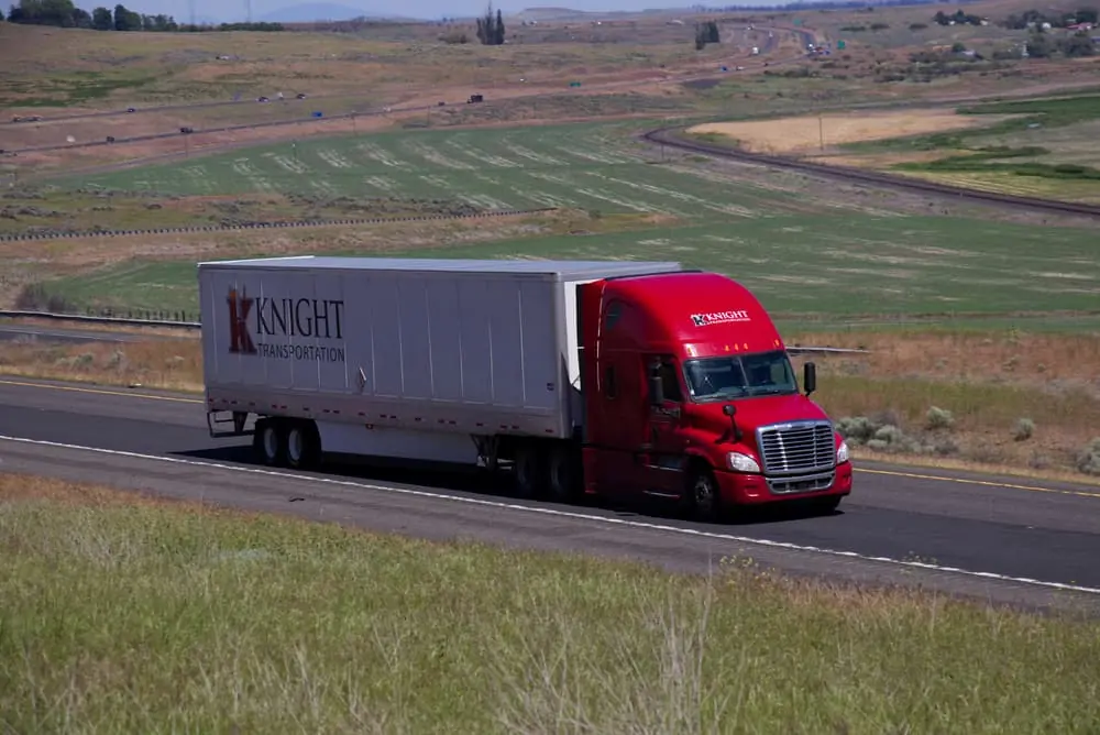 A Red Freightliner pulls a white trailer along a rural US Highway