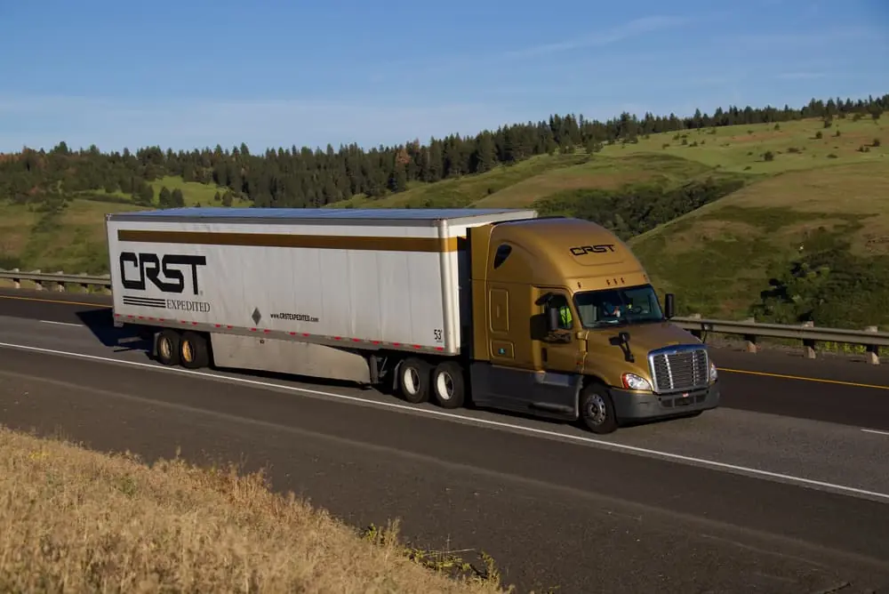 A gold freightliner pulls a white and gold trailer along a rural US Highway