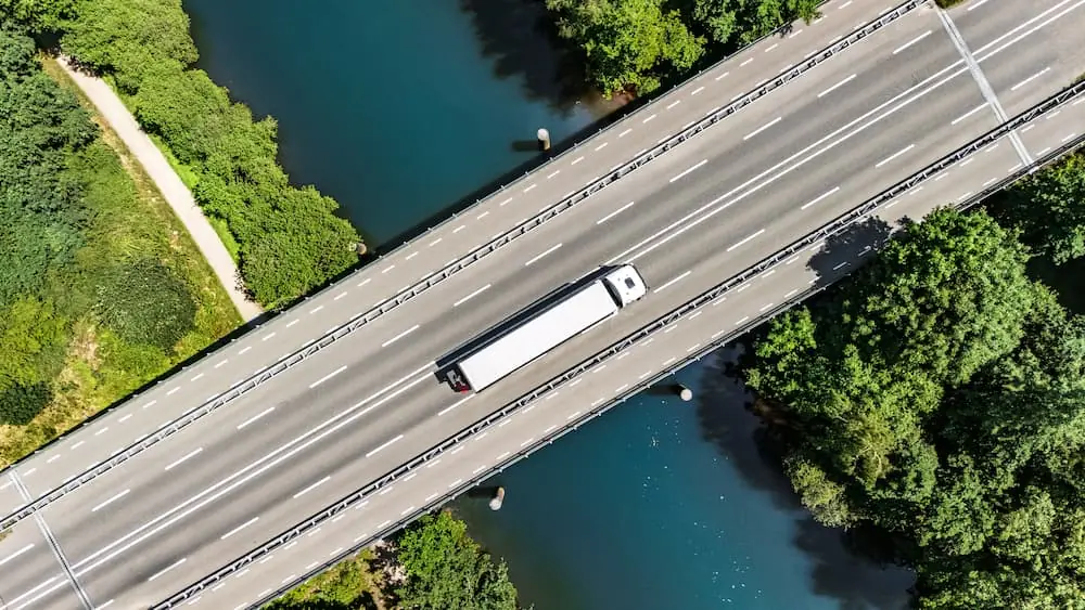 Freight Shipping in the Spring topdown aerial view of white semi truck driving across a bridge above a river during late spring