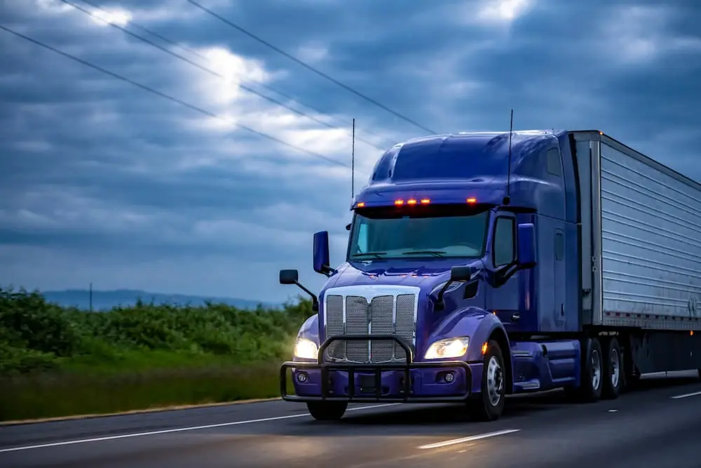 The Climate Forces Reshaping Freight in 2026 blue semi trailer driving with headlights on down rural highway at night