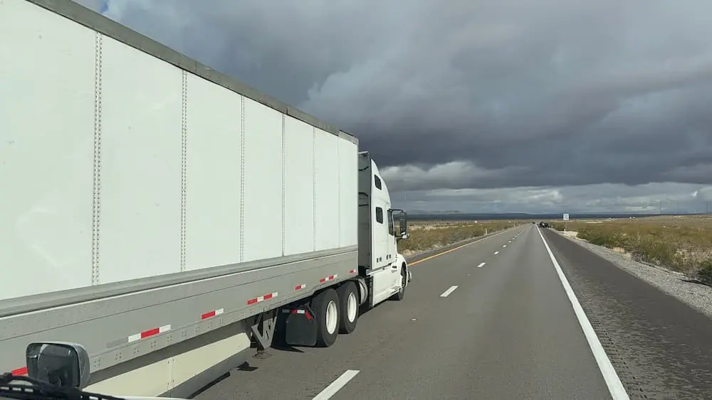 The Climate Forces Reshaping Freight in 2026 white semi trailer driving down desert highway as dark storm clouds gather above