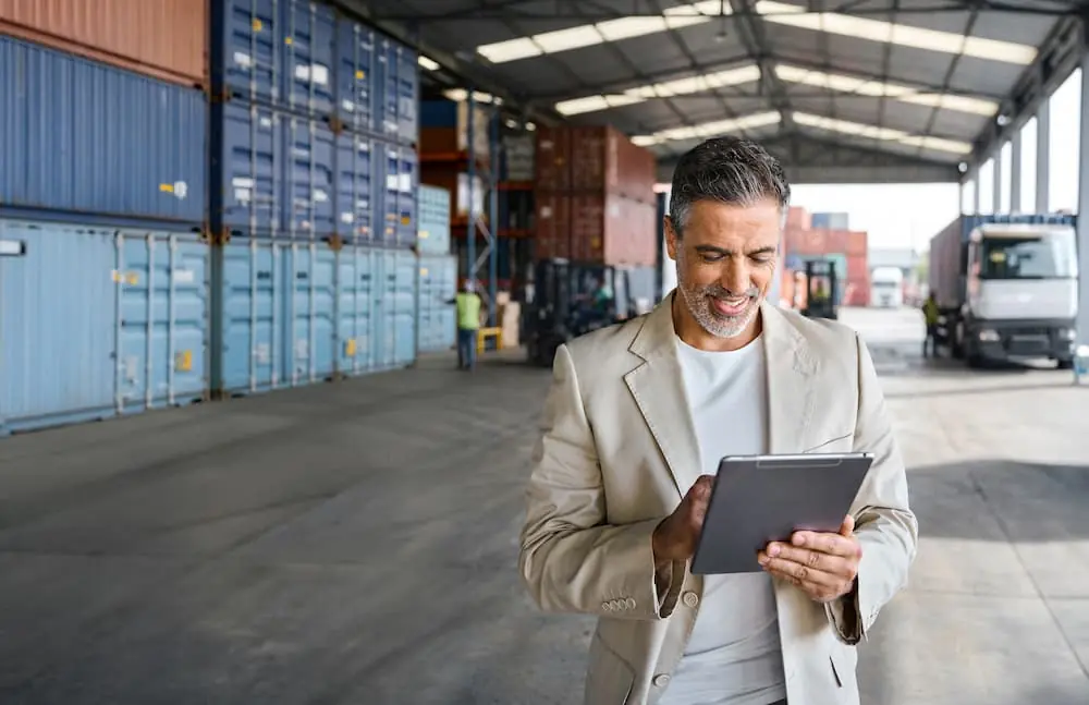 Logistics Talent Shortage logistics team lead looking over data on tablet while workers behind him move shipping containers
