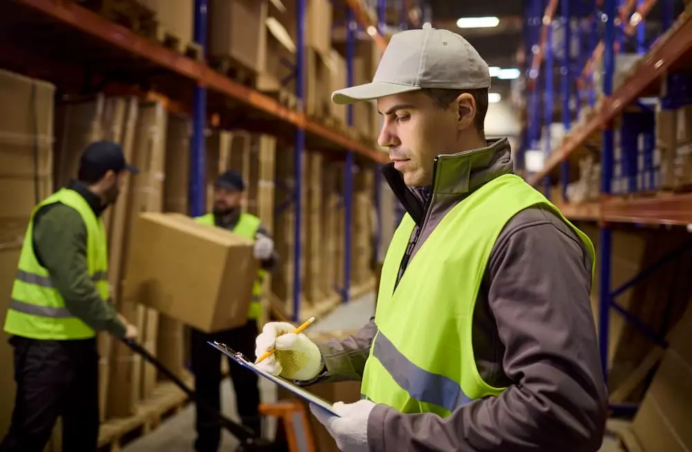 Logistics Talent Shortage warehouse worker looking over inventory data while workers behind him load cardboard boxes onto pallets