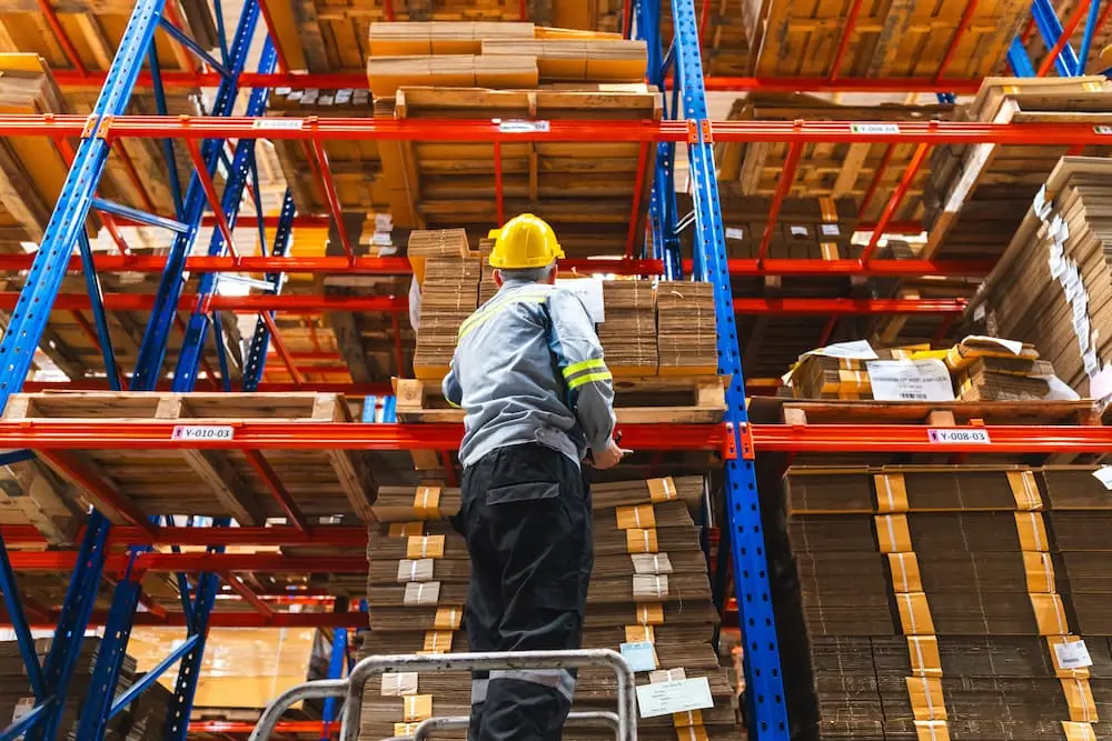 Logistics Talent Shortage warehouse worker on ladder looking up at warehouse shelf with clipboard in hand