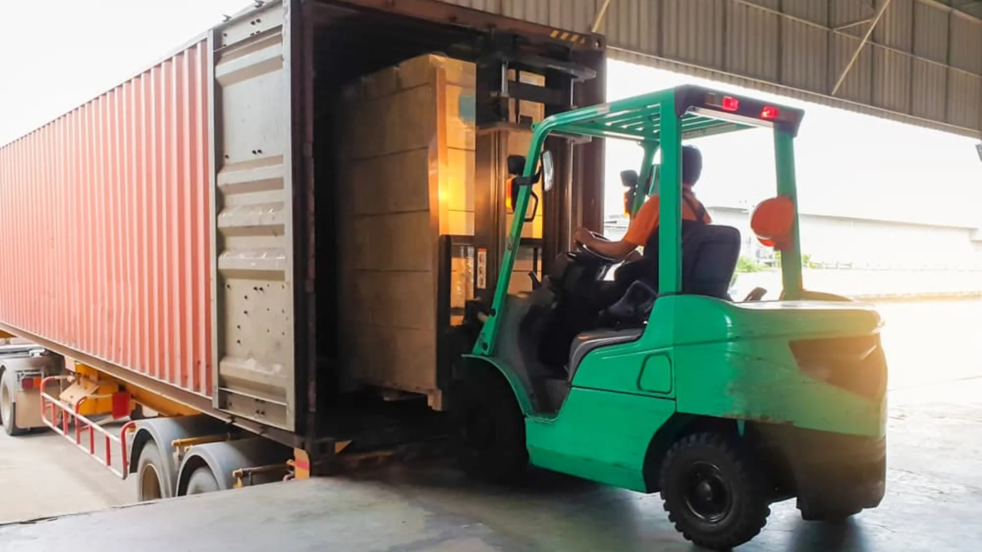 Forklift Tractor loading package boxes into shipping container at dock warehouse.