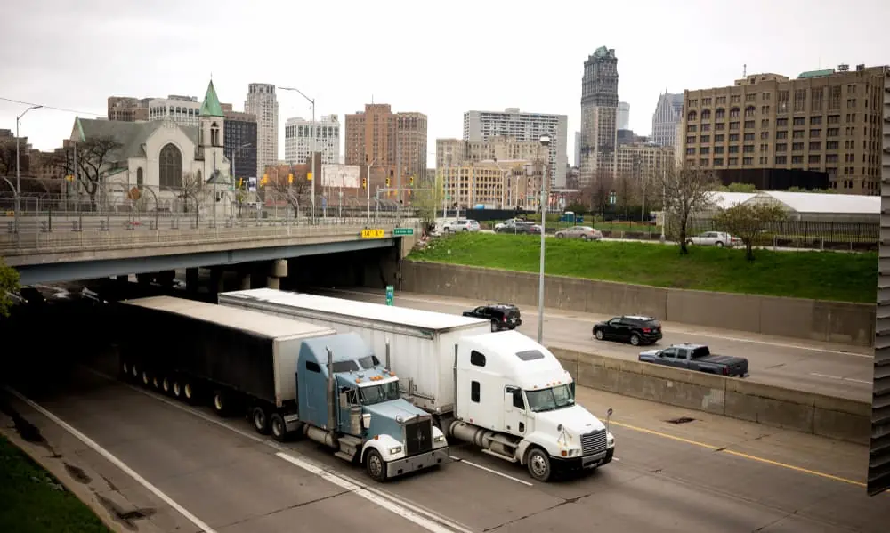 Truck Bottlenecks semi trailer trucks passing under interchange as cityscape looms behind them