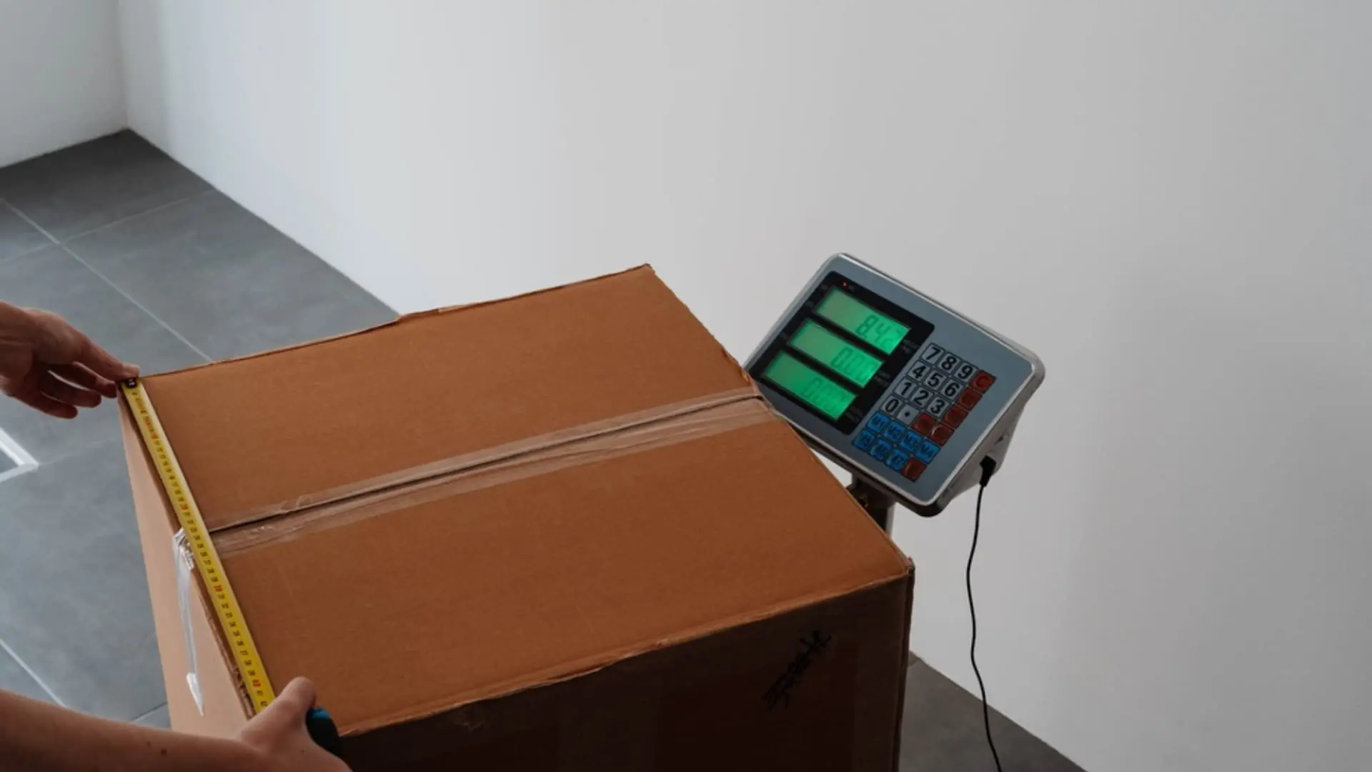 Hands stretch a tape measure along a sealed cardboard box resting on a digital platform scale.